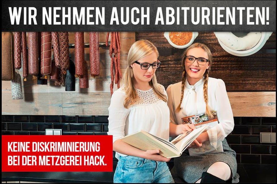 2 junge Frauen mit Brille und langen blonden Haaren. Eine schaut in ein Buch und eine lächelt in die Kamera. Letztere trägt einen kurzen Rock und schwarze Stiefel. Aufgenommen in den Fleischerei Hack und an der Wand hängen verschiedene Würste. Auf dem Bild ein Text: Keine Diskriminierung bei der Metzgerei Hack. Wir nehmen auch Abiturienten!