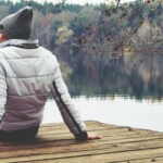 Mensch in herbstlicher Kleidung sitzt auf einem Holzsteg und schaut aufs Wasser. Im Hintergrund Bäume am Ufer des Sees. Die Szene steht für lebensphasenorientierte Personalarbeit und wirkt nachdenklich