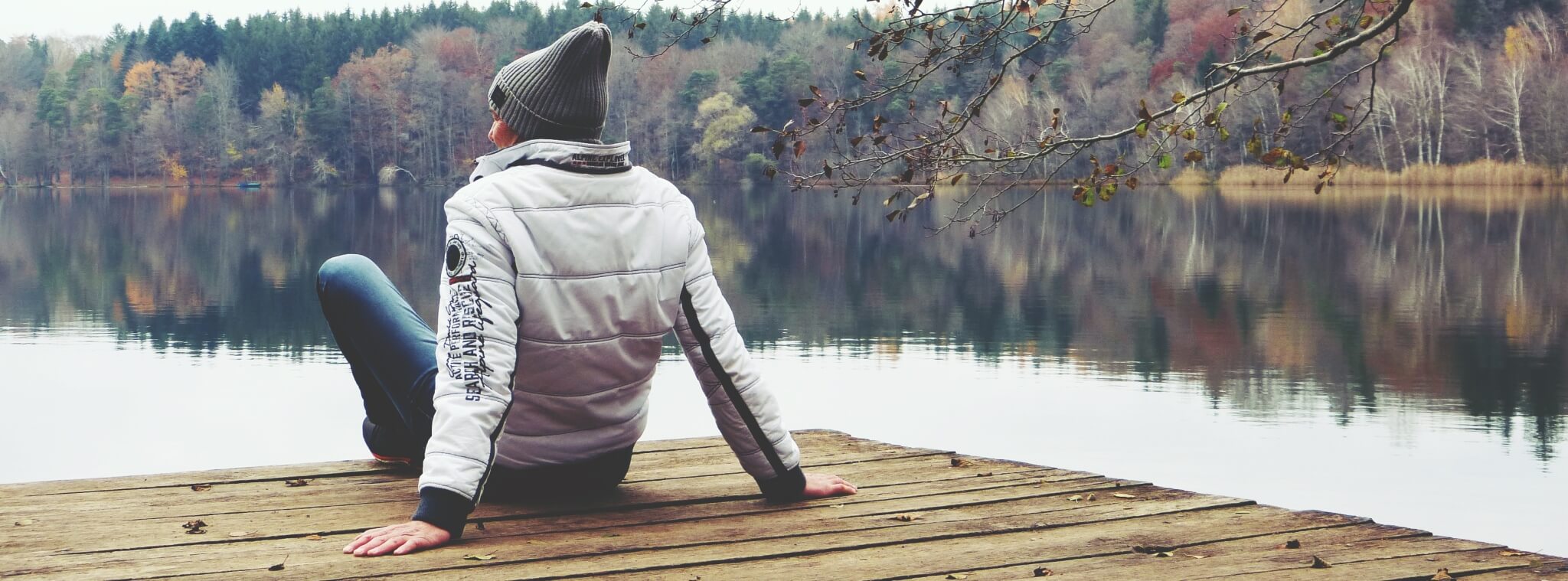Mensch in herbstlicher Kleidung sitzt auf einem Holzsteg und schaut aufs Wasser. Im Hintergrund Bäume am Ufer des Sees. Die Szene steht für lebensphasenorientierte Personalarbeit und wirkt nachdenklich